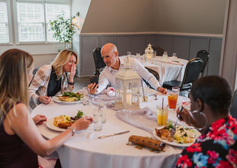 Four people seated at a table, eating breakfast, as two lean towards each other looking at a mobile phone.