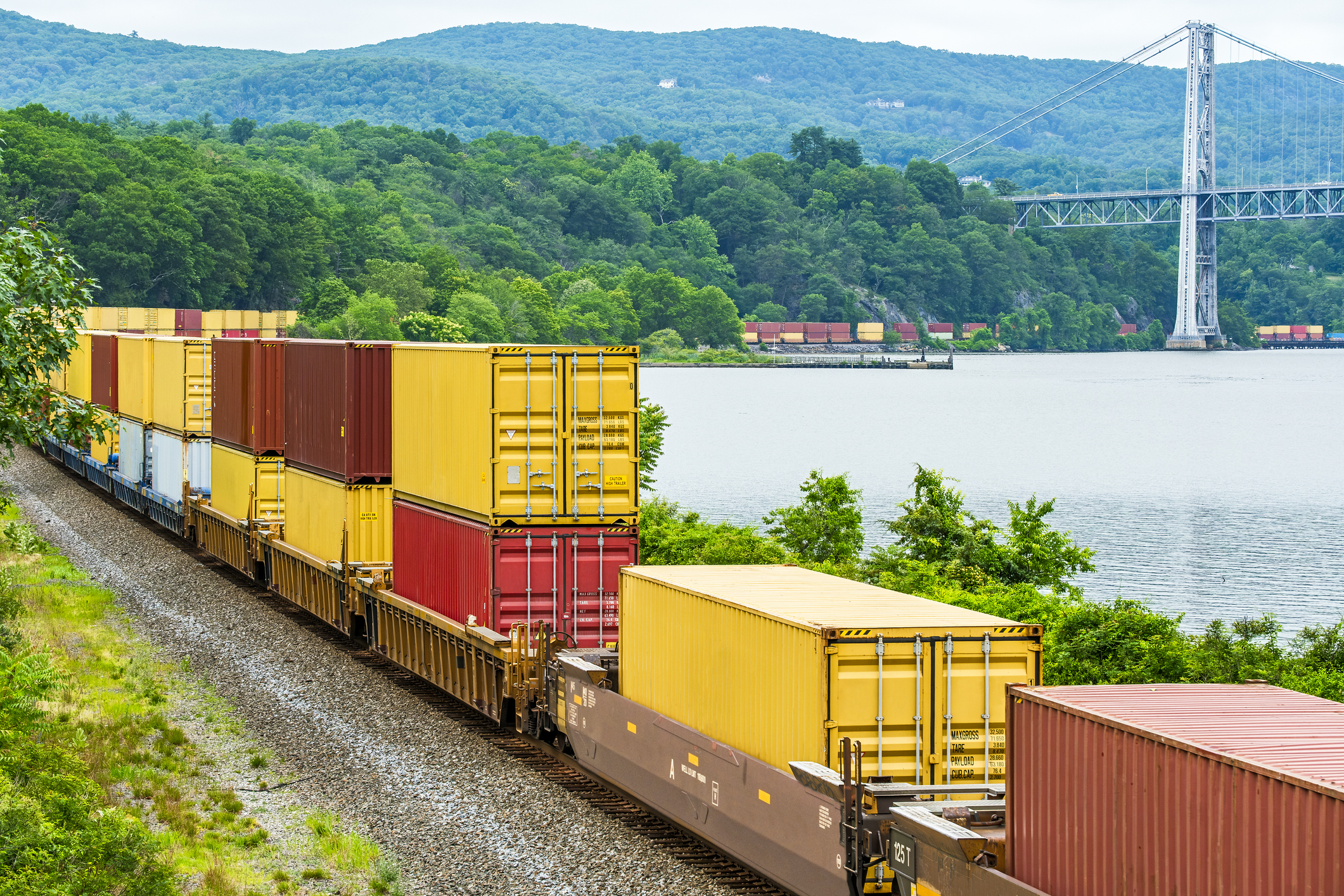 Container stack freight train rounding curve along Hudson River edge with Bear Mountain bridge